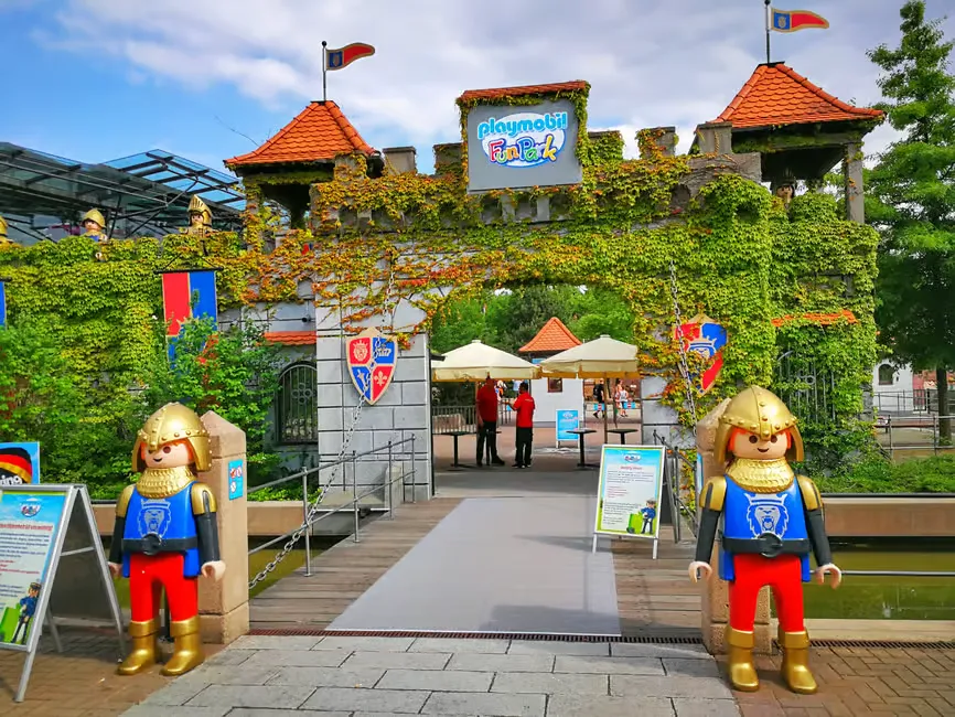 Two Playmobil figures dressed as knights stand by a castle entrance adorned with ivy. A colorful sign reads Playmobil FunPark.