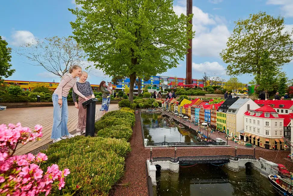 Kids observe a miniature town at a park, with colorful buildings and a small canal. Bright flowers and trees surround the scene.