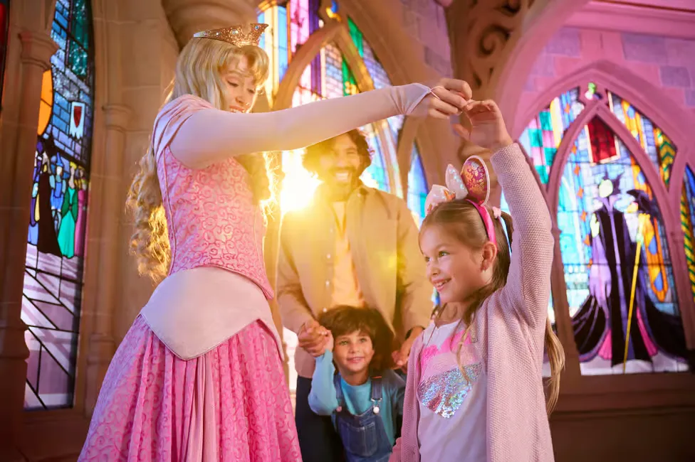 Princess in a pink gown twirls a smiling girl with Minnie ears in a castle setting. Stained-glass windows shine brightly behind them.