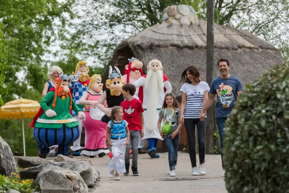 Families and children walk happily with cartoon characters at a theme park. Lush greenery and a thatched hut are in the background.