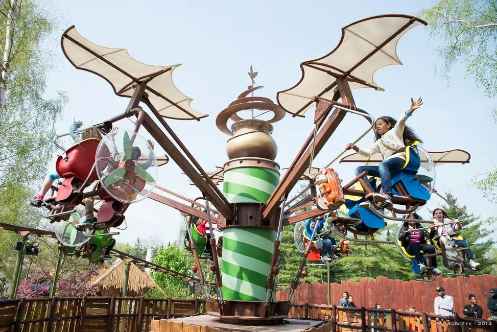 Kids ride a whimsical, colorful amusement park swing with dragonfly wings. Bright sunny day, trees and wooden fence in the background. Enthusiastic mood.