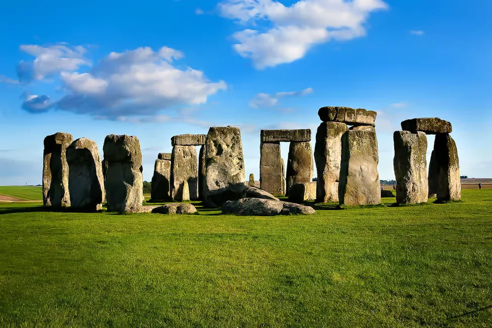 Stonehenge under a bright blue sky with scattered clouds, set on a green field. The ancient stone circle evokes a sense of mystery.