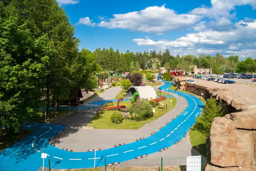 Aerial view of a park with a vibrant blue winding path, trees, and a Ferris wheel. Clear skies and parking lot in the background.