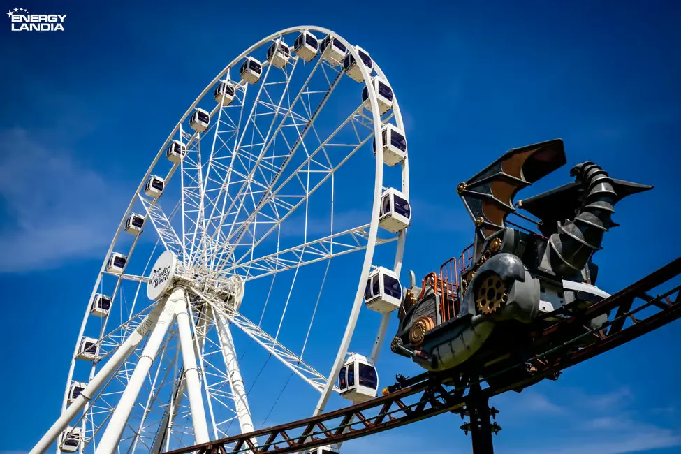 Ferris wheel next to dragon-themed roller coaster against a blue sky. "Energylandia" logo in the corner. Fun, exciting atmosphere.