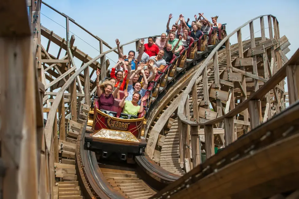 People on a wooden roller coaster raise their hands, showing excitement. The coaster car is labeled "Wodan." Sunny day with a blue sky.