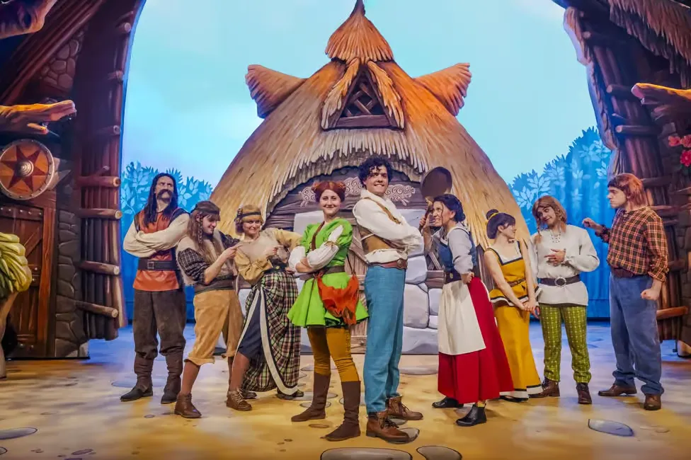 Performers dressed in colorful medieval costumes stand proudly on stage in front of a whimsical cottage backdrop.