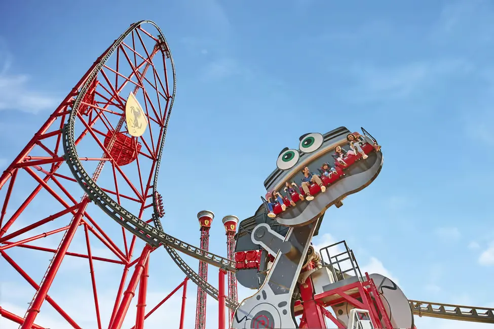 People ride a roller coaster with a cartoonish face at an amusement park. Bright red tracks loop against a clear blue sky.