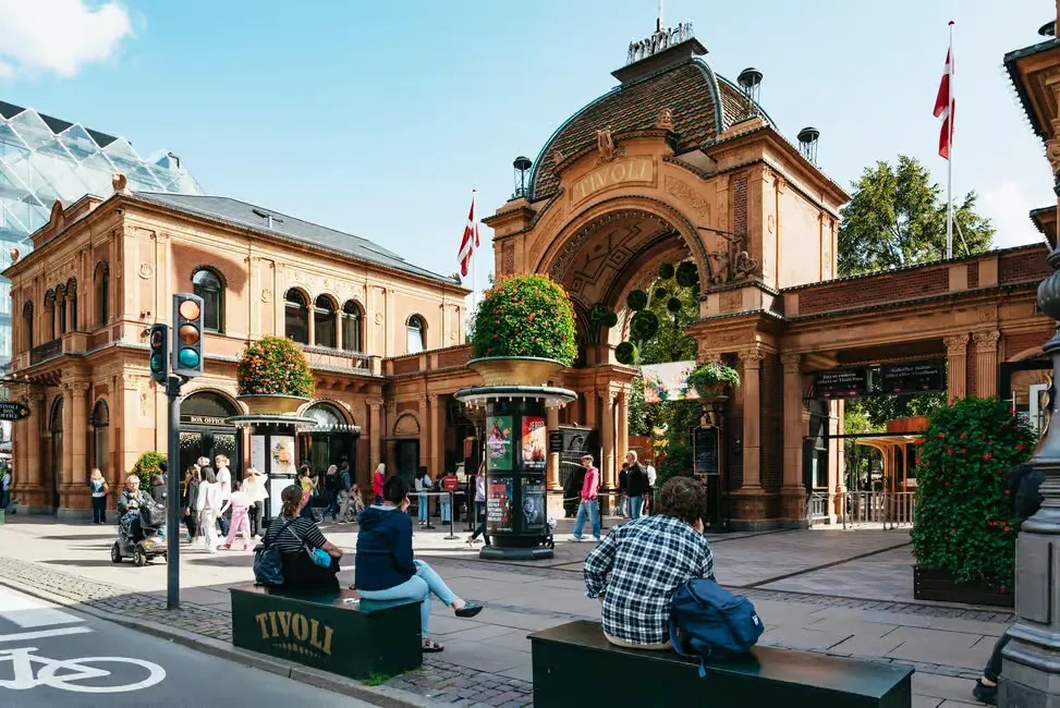 People walk by and sit near the entrance of Tivoli Gardens in Copenhagen. The ornate arch is flanked by Danish flags and greenery.