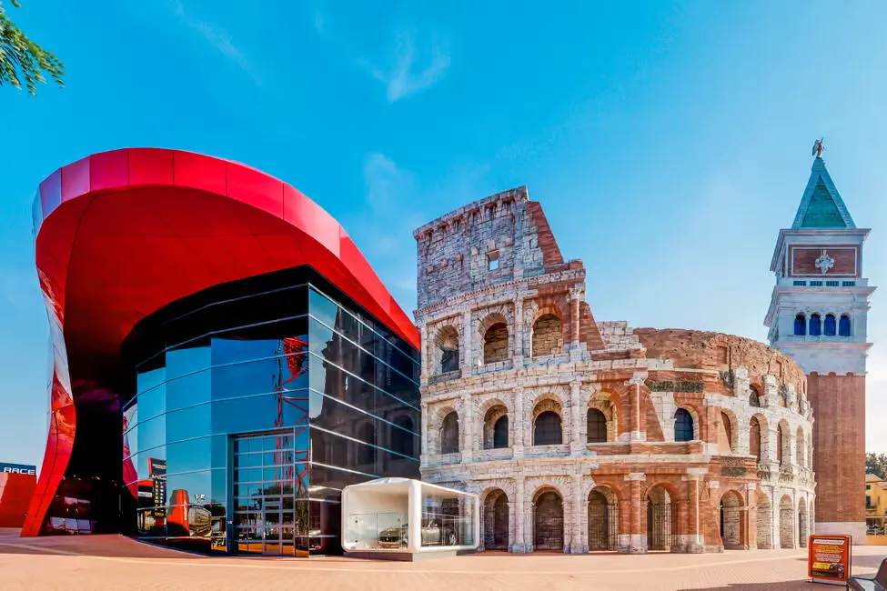 Modern red building beside a Colosseum replica and bell tower under a clear blue sky. Glass windows reflect bright daylight.