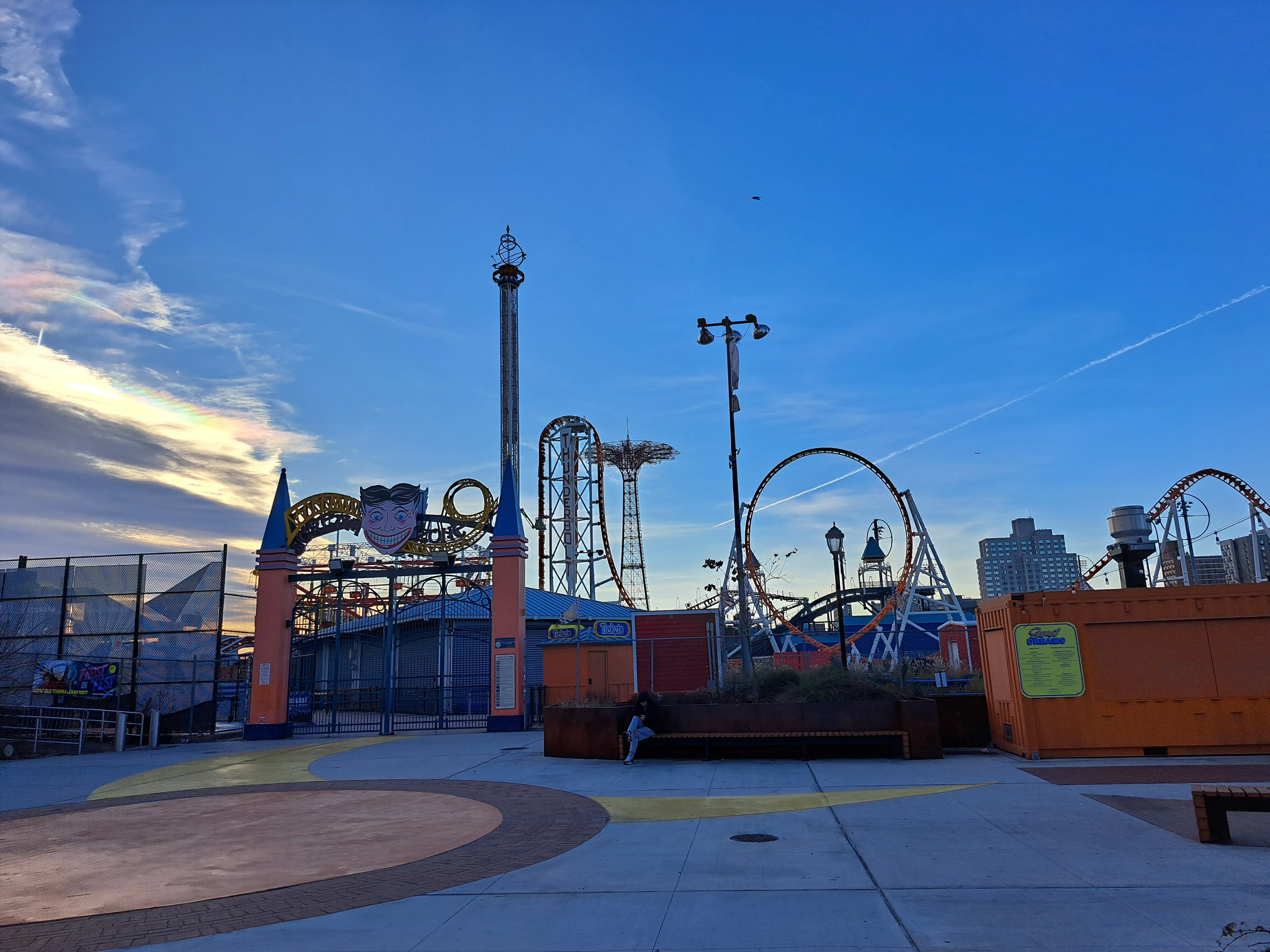 Amusement park entrance with roller coasters against a clear blue sky. Orange storage unit on the right with some visible text.