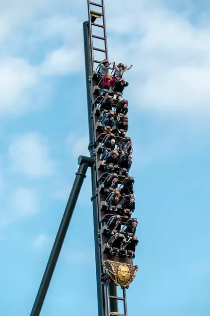 People ride a vertical roller coaster against a blue sky, some with raised arms, creating an atmosphere of thrill and excitement.