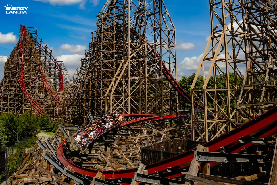 Roller coaster with wooden structure and red tracks at Energylandia. People riding with excitement amid trees and blue sky background.