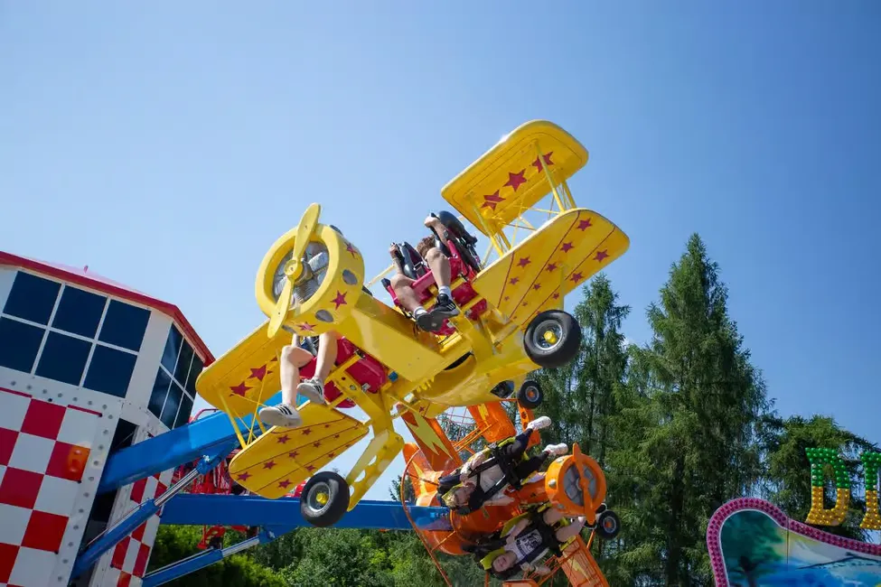 Bright yellow and orange airplane ride with people enjoying, under a clear blue sky, near trees and a checker-patterned building.