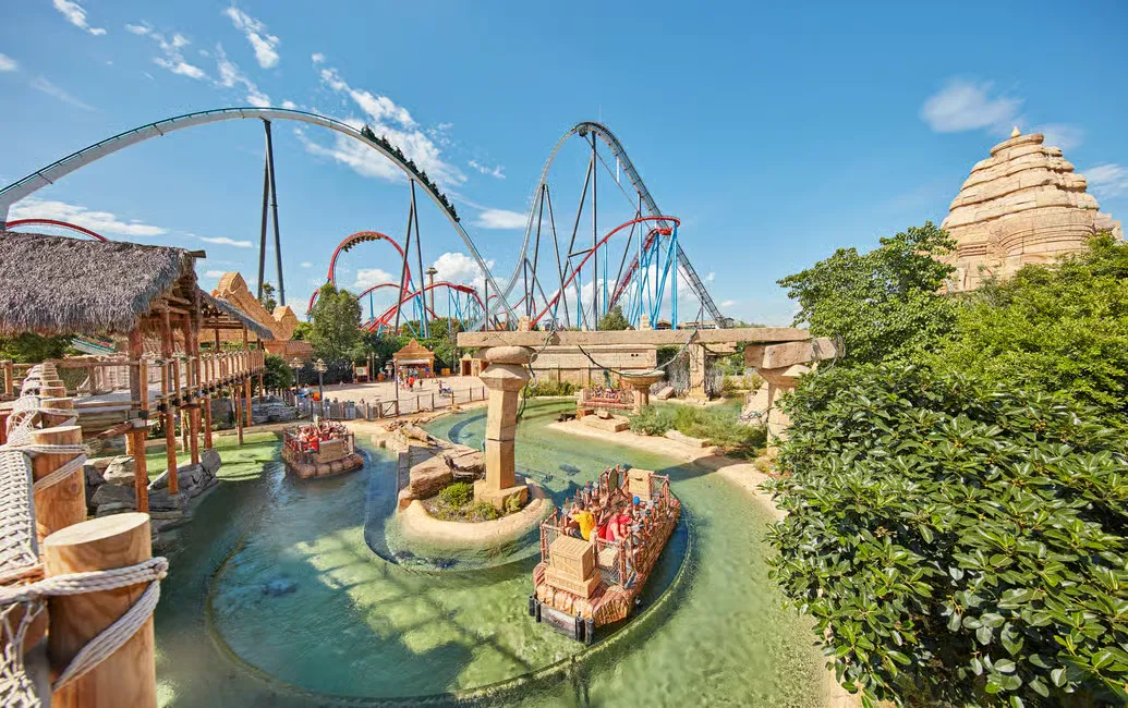 Theme park with vibrant roller coasters in the background. People enjoy a boat ride on clear water. Sunny day with blue skies.