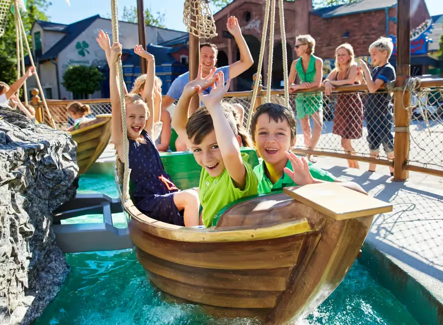 Children joyfully raise hands on a swinging boat ride, surrounded by water. Onlookers watch from behind a fence. Sunny day, vibrant colors.