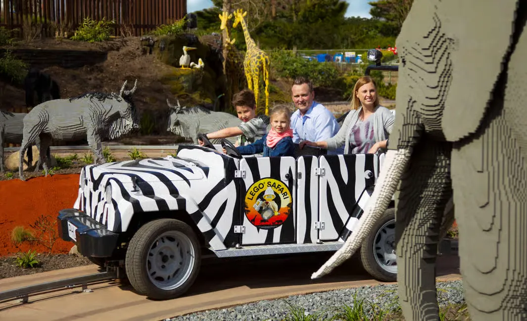 Family on zebra-striped LEGO Safari vehicle, surrounded by LEGO animals like elephants and giraffes. Bright, cheerful outdoor setting.