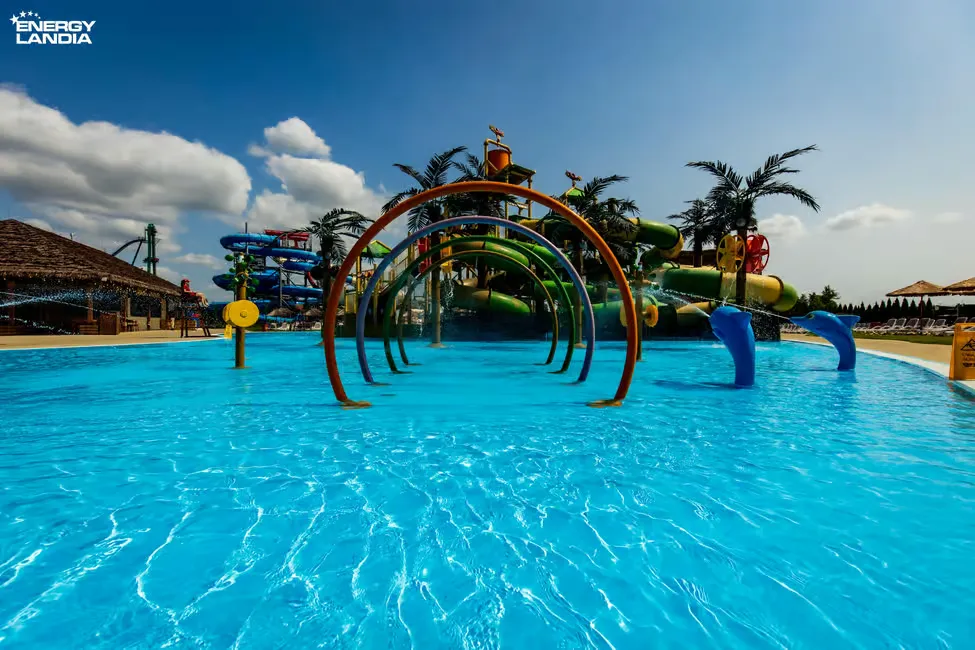 Water park scene with colorful slides and arches spraying water into a bright blue pool under a clear sky. "Energylandia" text visible.
