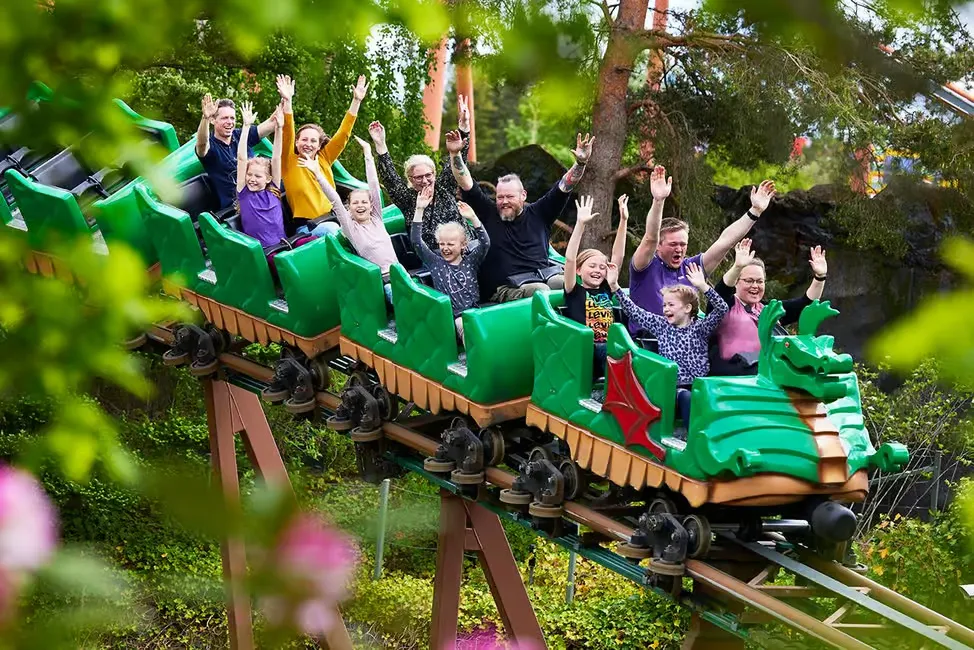 People on a green dragon-themed roller coaster, raising their arms, smiling. The ride is surrounded by lush greenery, creating a joyful vibe.
