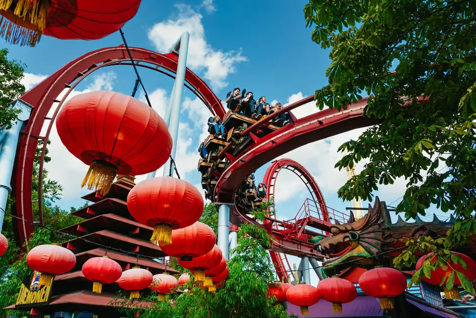 People on red roller coaster under blue sky with red lanterns and dragon decoration. Trees surround the vibrant, festive setting.