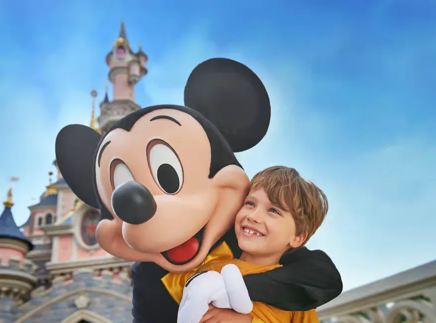 Child hugging Mickey Mouse in front of a colorful castle. Blue sky background. The child is smiling, creating a joyful atmosphere.