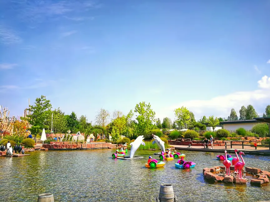 Pond with colorful sculptures of flamingos and dolphins, surrounded by lush greenery. People walking in the background under a clear blue sky.