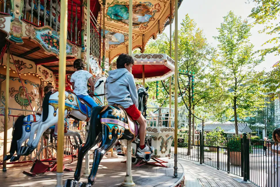 Children ride painted horses on a colorful carousel in a park. Bright patterns decorate the ride, surrounded by green trees.