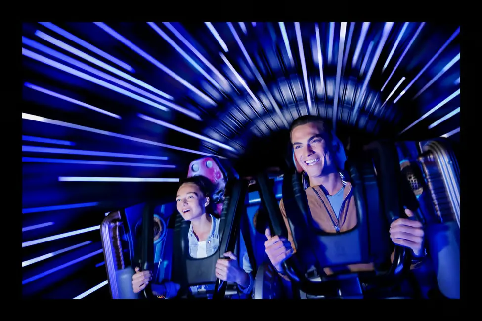 Two people on a roller coaster, smiling with excitement. The background is a blur of blue lights, creating a fast-paced, thrilling atmosphere.