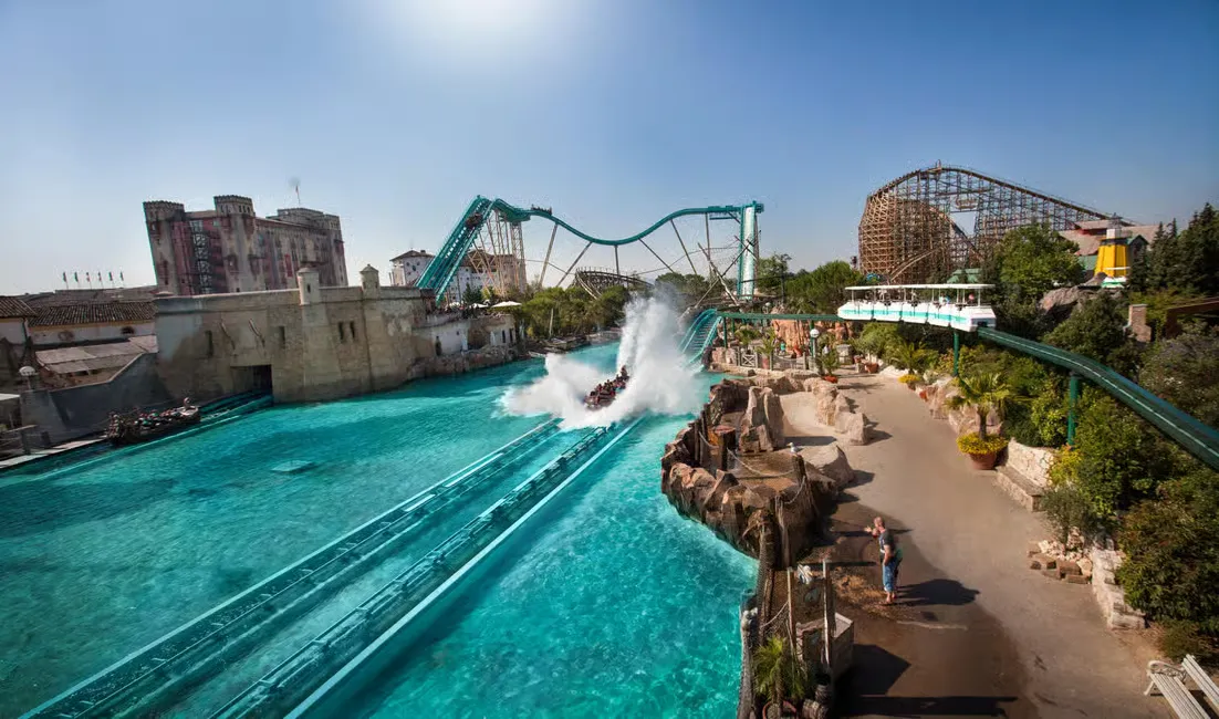 Roller coaster drops into bright blue water, creating a huge splash. Amusement park with rides and people on path, sunny day.