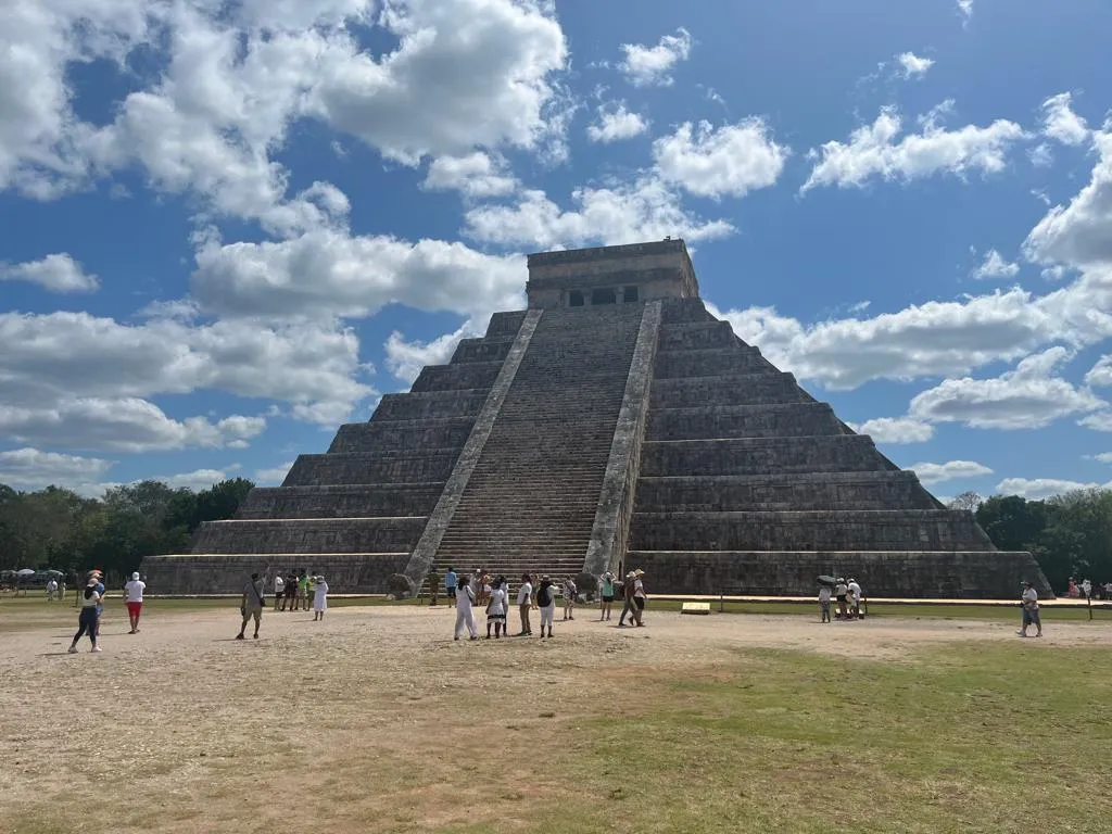 People gather around the stone pyramid at Chichen Itza under a partly cloudy blue sky, creating a lively, historic atmosphere.
