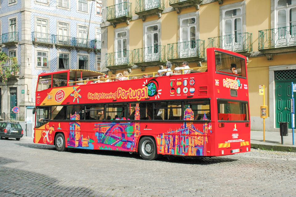 Red double-decker sightseeing bus with colorful illustrations tours near traditional Portuguese buildings. Passengers enjoy the open top. Text: "CitySightseeing Portugal".