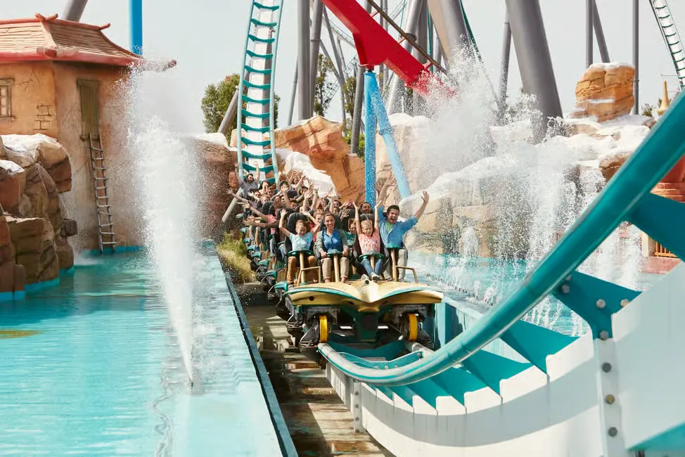 People riding a roller coaster through water splashes, raising hands and smiling. Blue tracks, rocky background, sunny day, joyful mood.