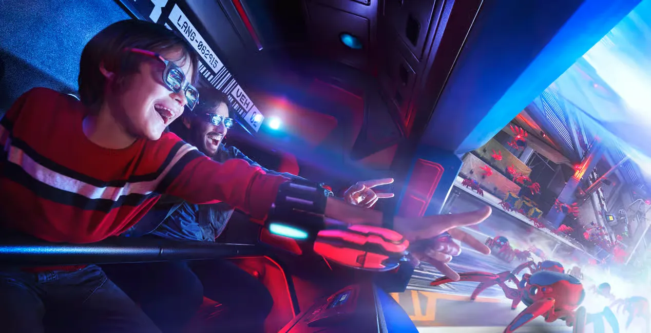 Child and adult wearing 3D glasses, excitedly pointing at animated red spiders on a screen. Bright, colorful lighting surrounds them.