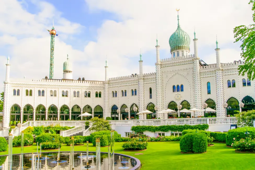 Ornate white building with a green dome, surrounded by lush gardens and a reflective pond. Bright, clear sky in the background.