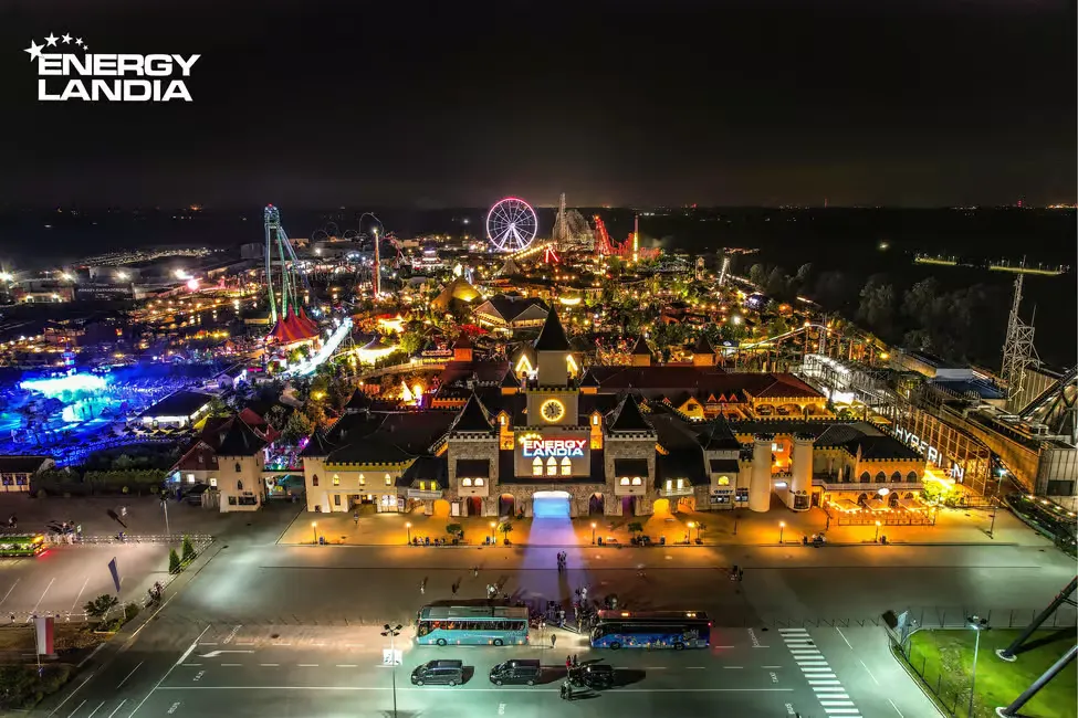 Nighttime aerial view of a brightly lit amusement park with rides, a Ferris wheel, and colorful lights; Energylandia logo on a building.