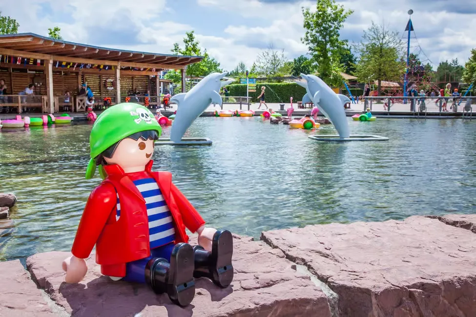 Toy pirate figure in red coat and green hat sits by a pond with dolphin sculptures. Colorful floats and people in the background.