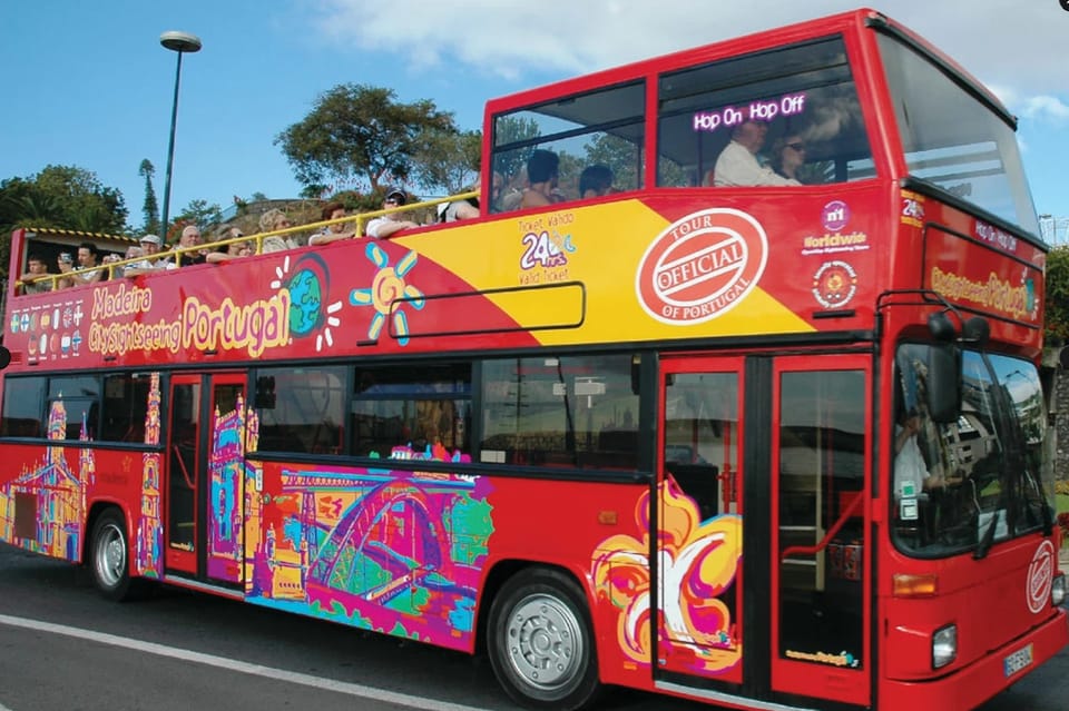 Red double-decker bus with tourists on top deck. Vibrant patterns, "Madeira City Sightseeing Portugal" text. Sunny day, trees in background.