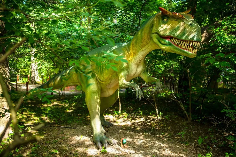 A large, green dinosaur statue with sharp teeth stands in a lush, sunlit forest setting, surrounded by trees and foliage.